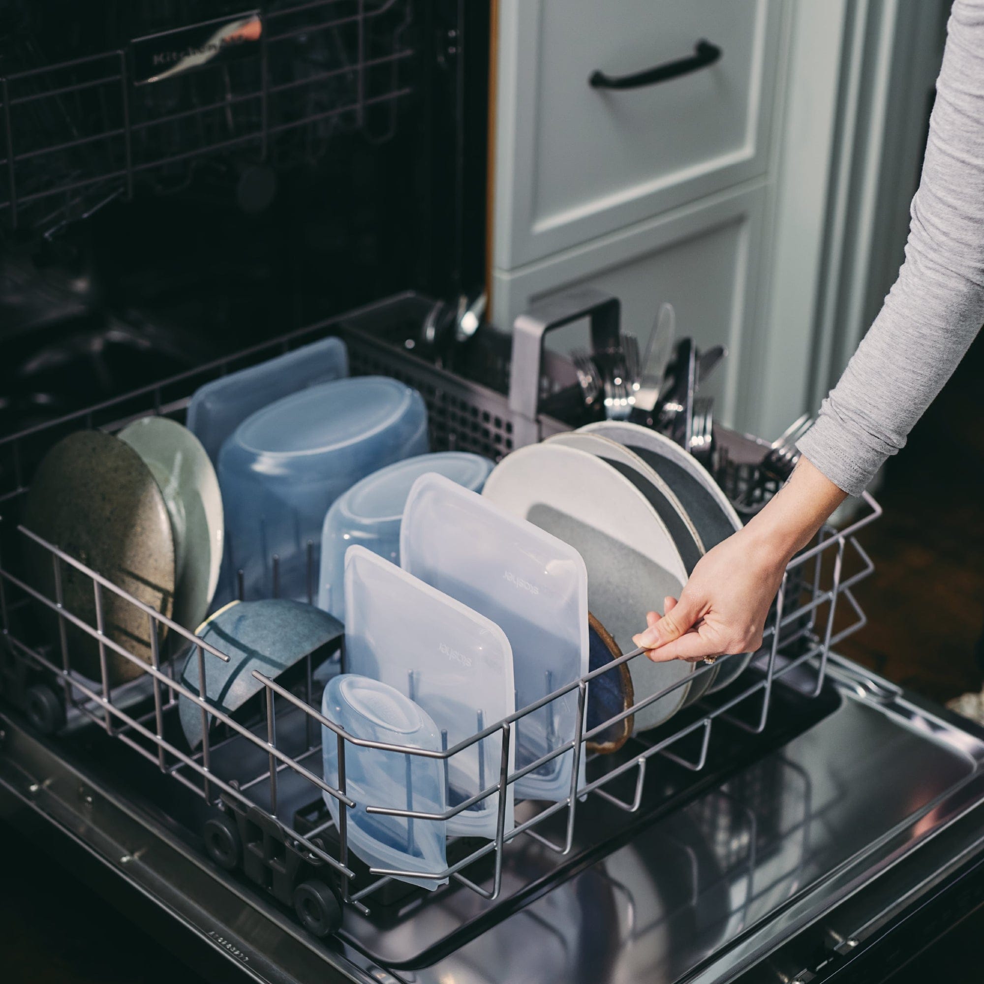 stasher bags and bowls in the bottom rack of a dishwasher being closd by a woman. 