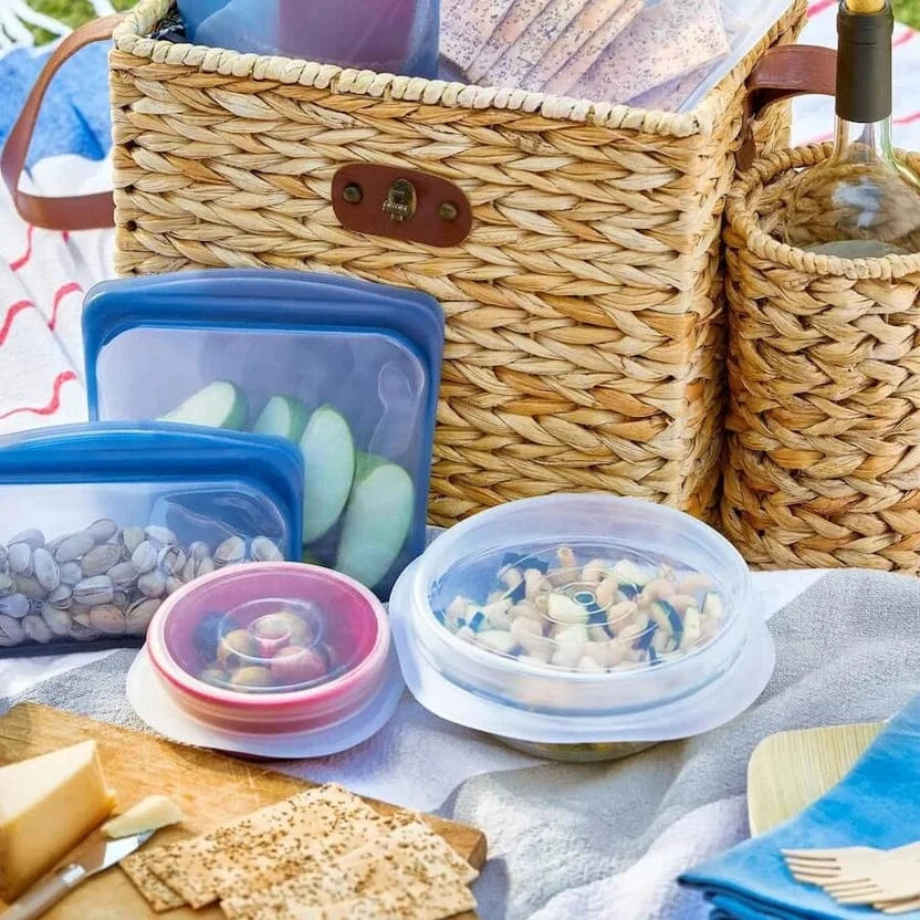 stasher bags and stretch lids being used at a picnic, holding sliced apples and pistachios and covering bowls of pasta salad.